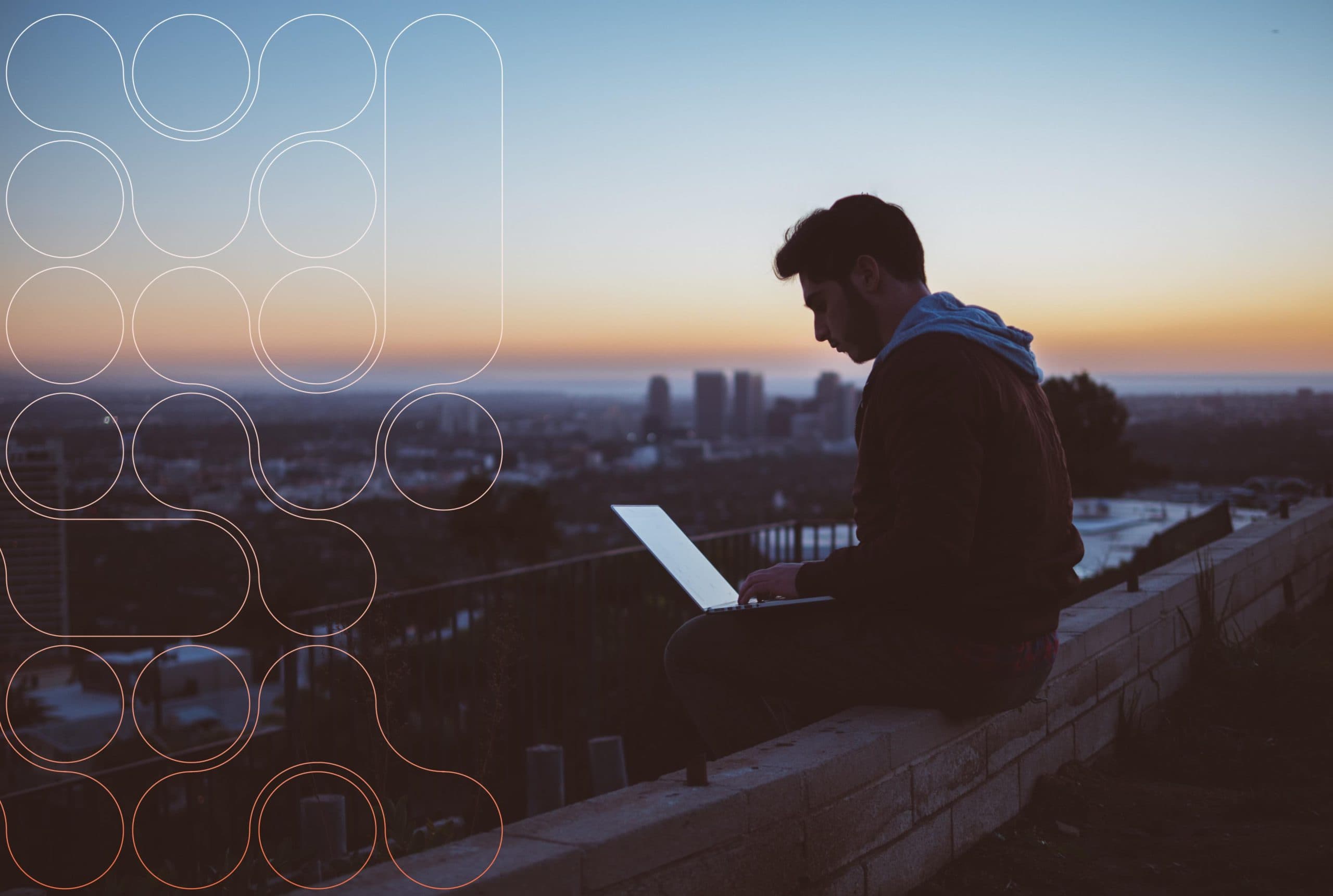 A photograph of a person sitting with a laptop with the city skyline and sunset in the background.