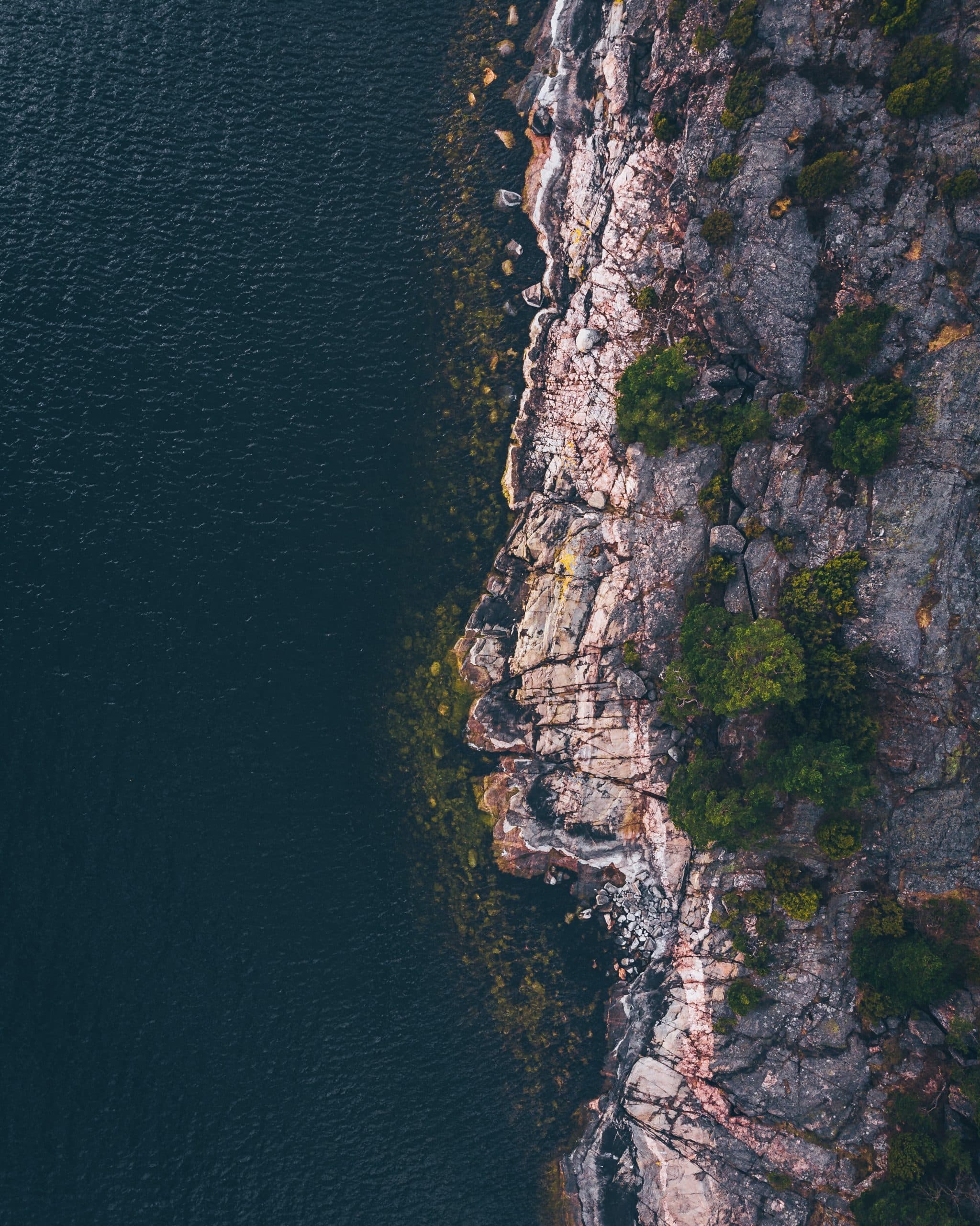 An aerial image taken top down over the swedish coast. On the left side there's water and on the right there's cliffs and a few trees.